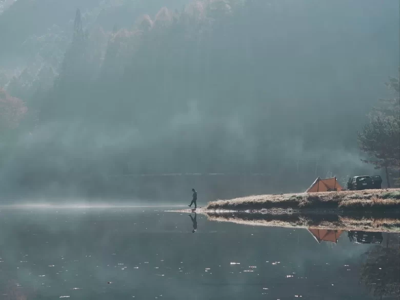 明け方の冷えて霧がかった湖の傍でキャンプをしている人